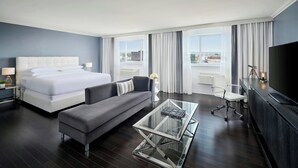 A modern hotel room with a large bed, a sofa, a glass coffee table, a desk with a flat-screen TV, and a view of the city through large windows.