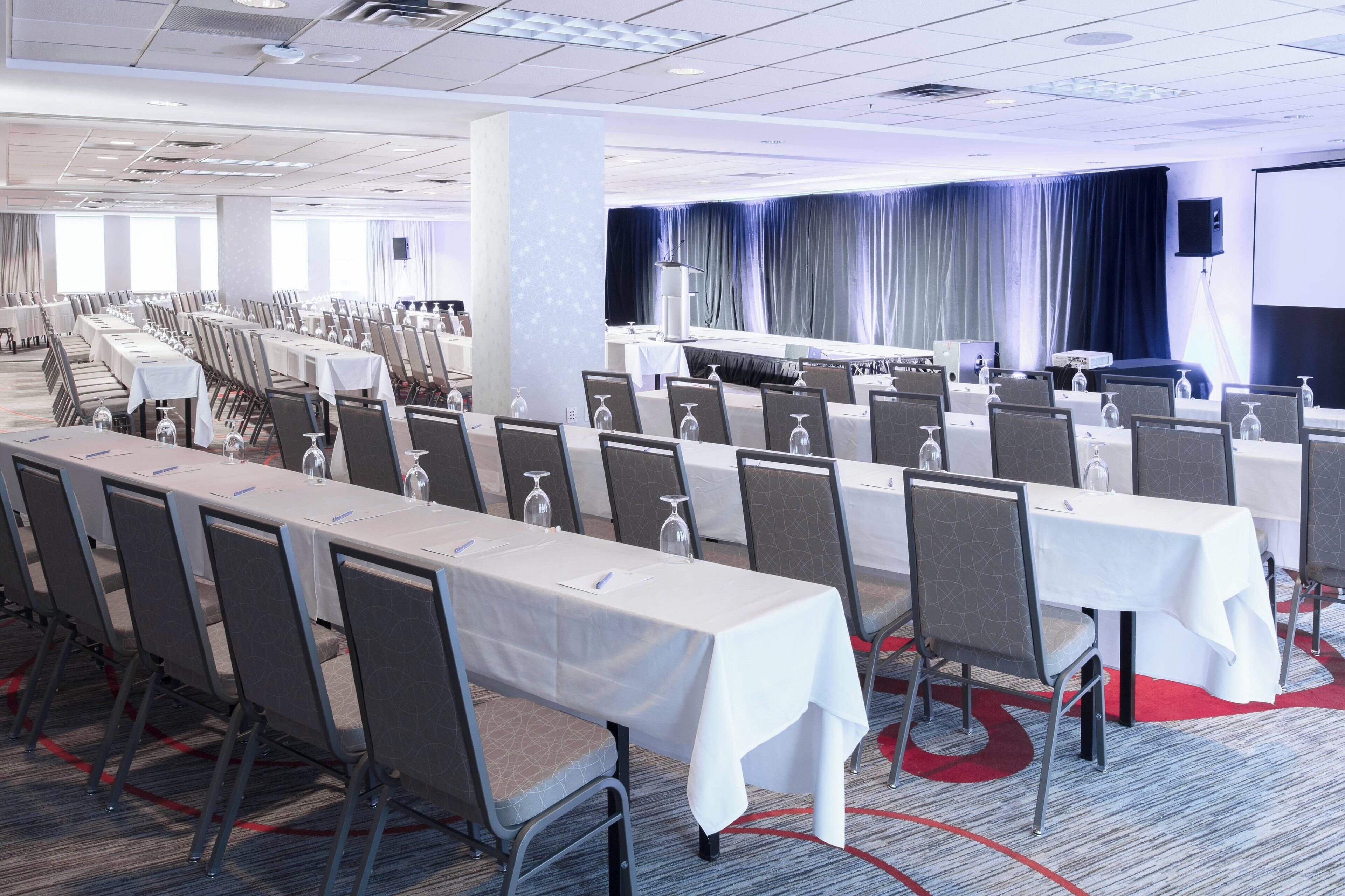 Un salón de conferencias con filas de mesas y sillas, un escenario con cortinas y una pantalla de proyector.