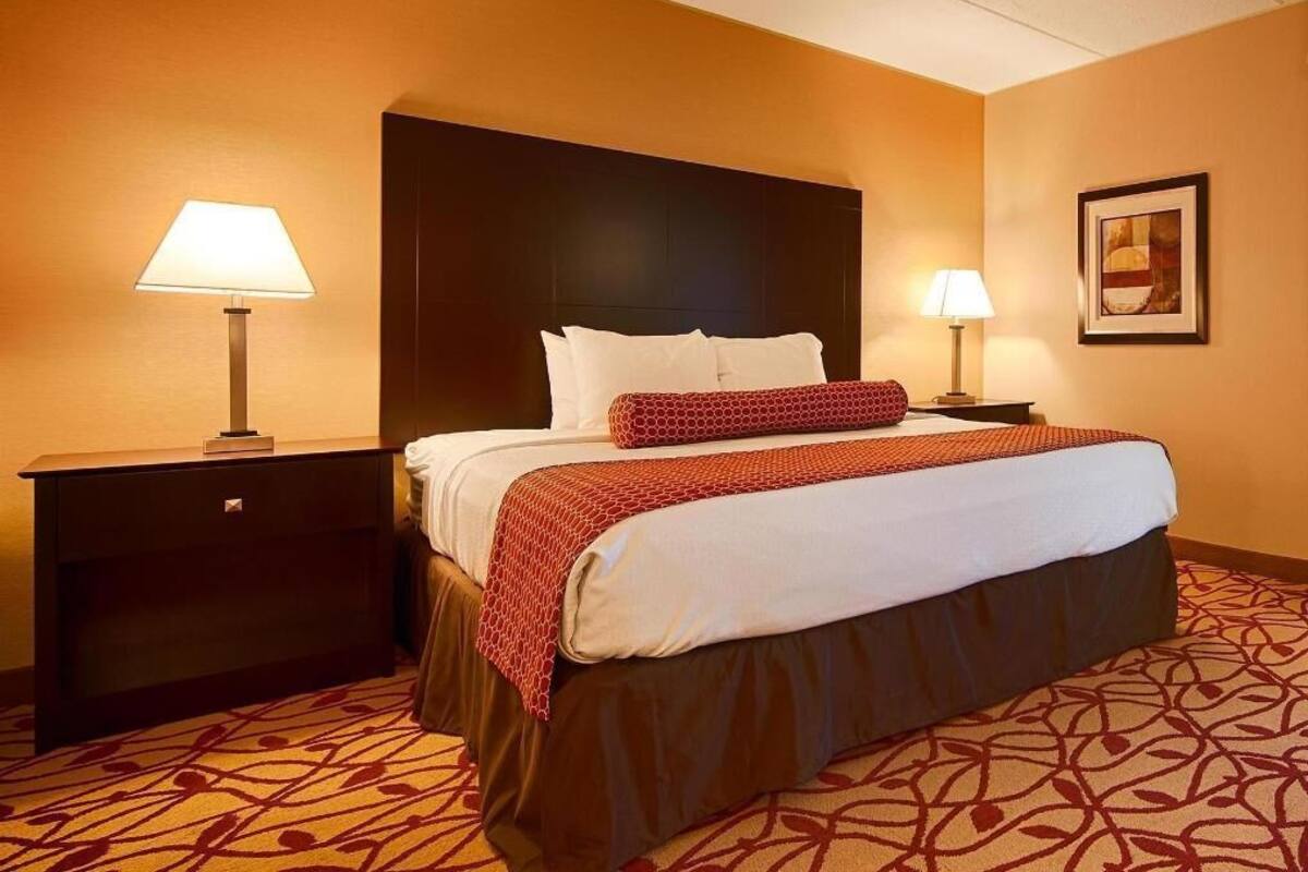 A hotel room with a large bed, two bedside tables, a headboard, a patterned carpet, and a framed picture on the wall.