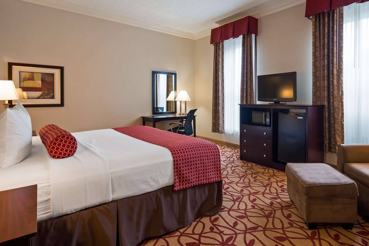 A hotel room with a bed, a desk with a chair, a television, a minibar, and a sofa.