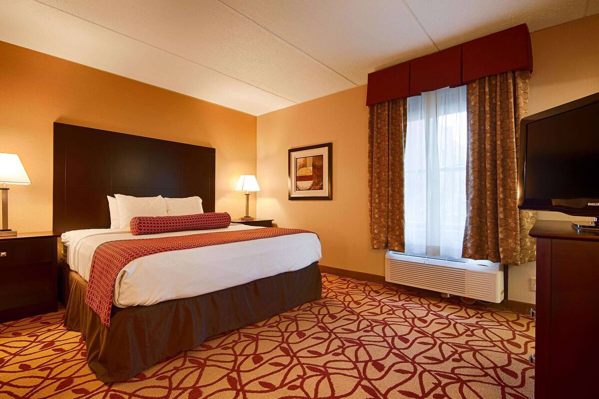 A hotel room with a bed, two bedside lamps, a flat-screen TV, a window with curtains, and a wall-mounted air conditioner.