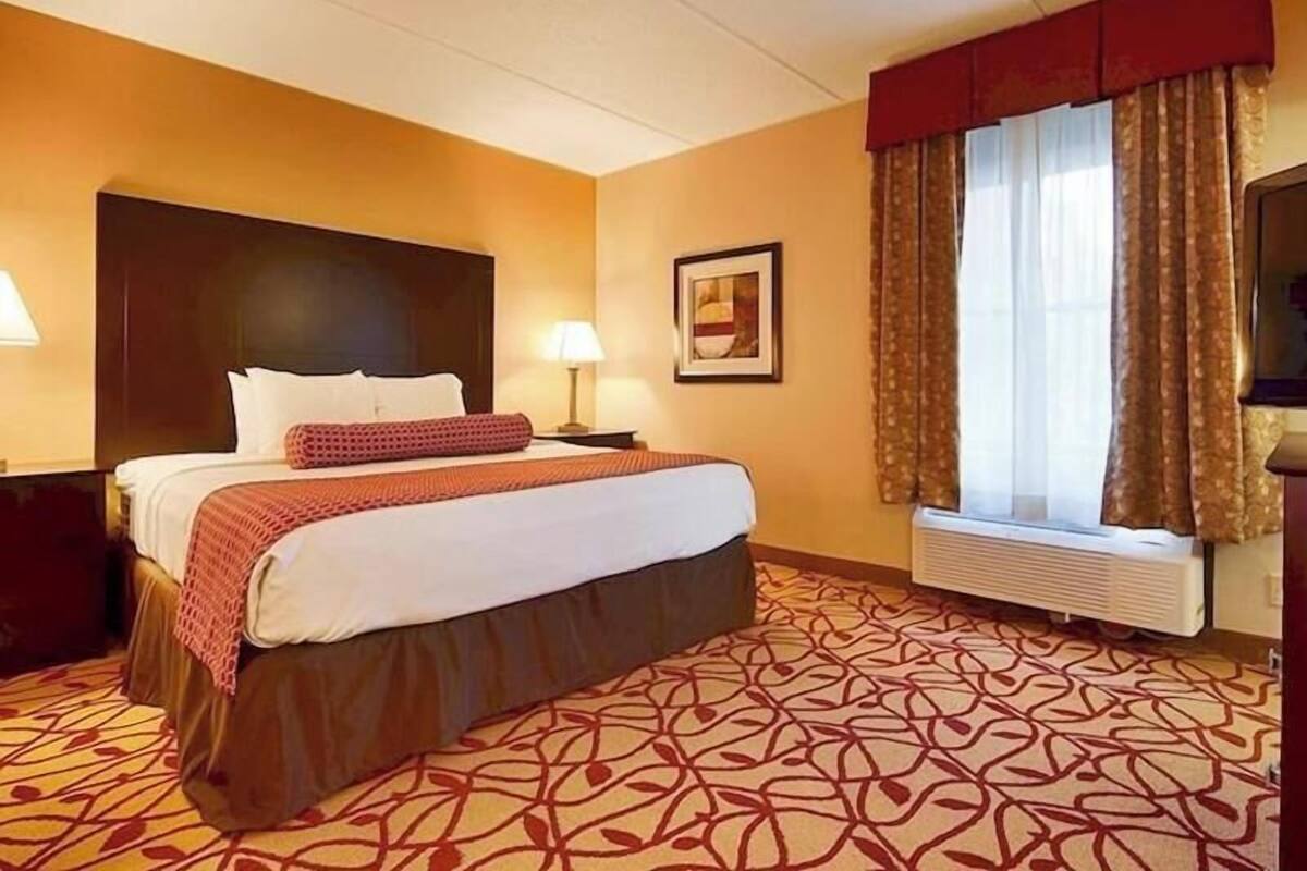 A hotel room with a bed, a nightstand, a lamp, a window with curtains, and a radiator.