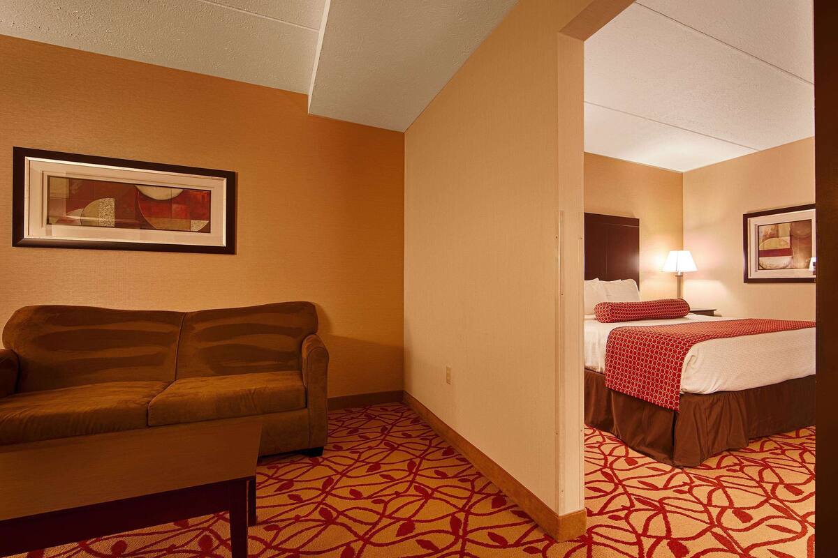 A hotel room with a sofa, a bed with a red and white patterned bedspread, a framed picture on the wall, and a patterned carpet.