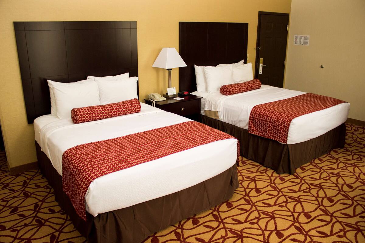 A hotel room with two beds, each with a brown skirted base and a red and white patterned bedspread. There is a nightstand with a lamp between the beds.