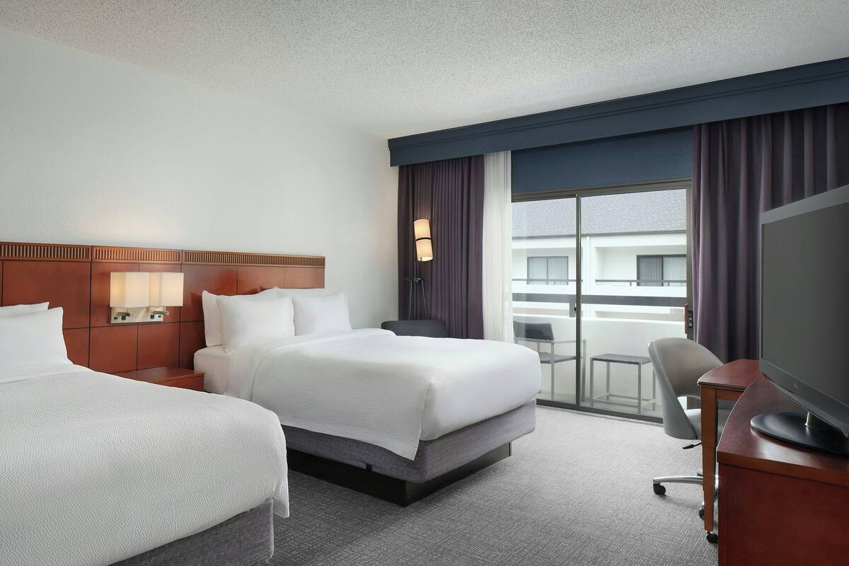 A hotel room with two beds, a desk with a computer, a TV, and a balcony with a view of buildings.