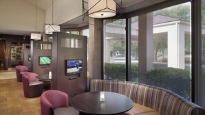 A modern hotel lobby with a seating area featuring a round table and cushioned benches, a television mounted on a partition, and large windows offering a view of the outside. - Courtyard Charlotte Arrowood (Charlotte)