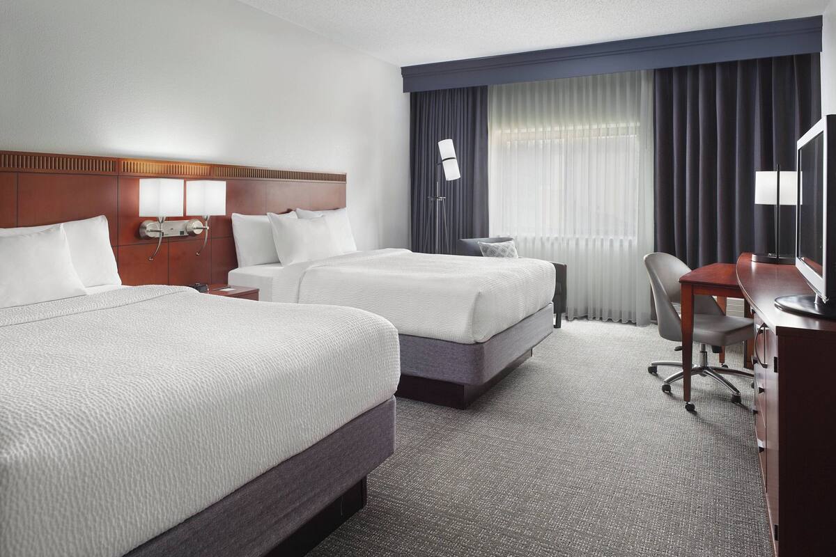A hotel room with two beds, a desk, a chair, a TV, and a window with curtains.