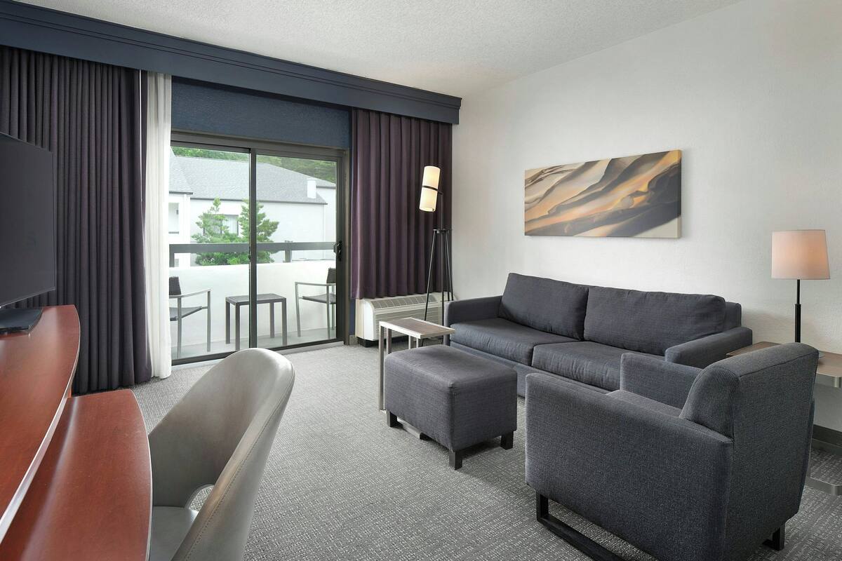 A modern hotel room with a grey sofa set, a wooden desk, a flat-screen TV, and a large window with curtains.