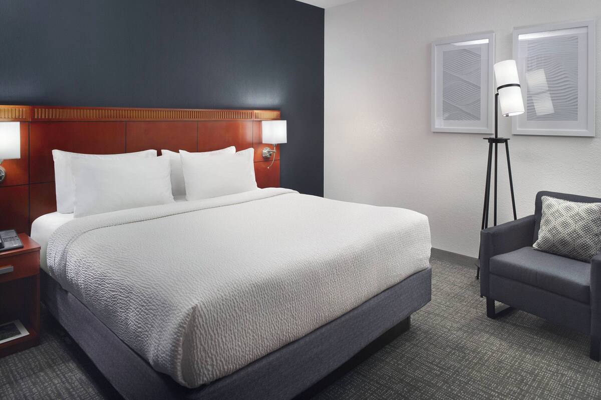A hotel room with a large bed, a bedside table with a lamp, a chair, and two framed pictures on the wall.