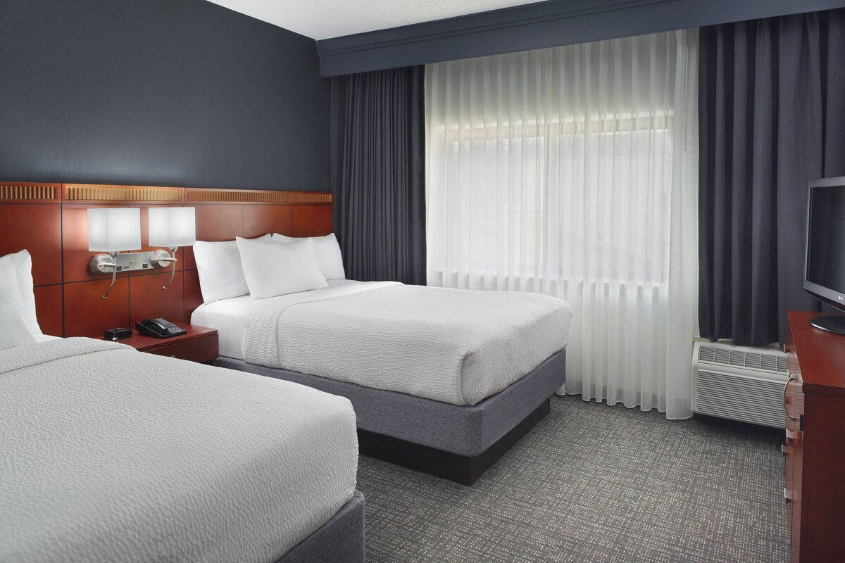 A hotel room with two beds, a wooden headboard, a bedside table with a phone, a TV, and a large window with curtains.