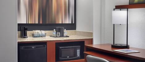 A compact hotel room with a kitchenette, a microwave, a coffee maker, and a desk with a lamp.