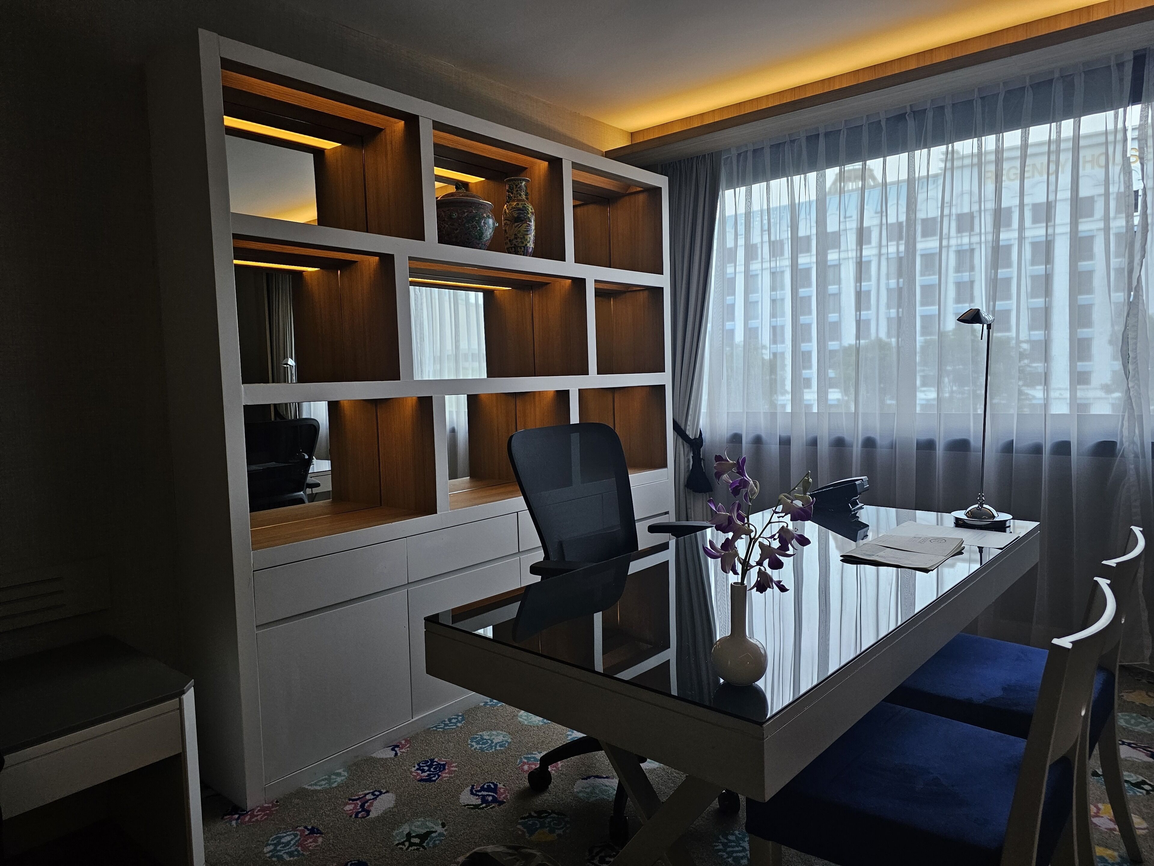 A modern office space with a glass desk, a black office chair, and a built-in shelving unit with decorative items.