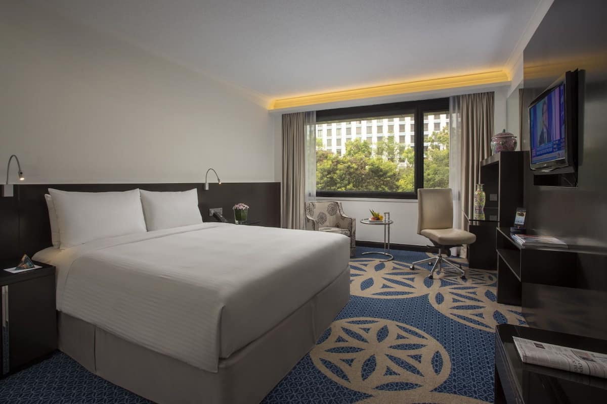 A hotel room with a large bed, a desk with a lamp, a chair, a TV, and a window with a view of buildings and trees.