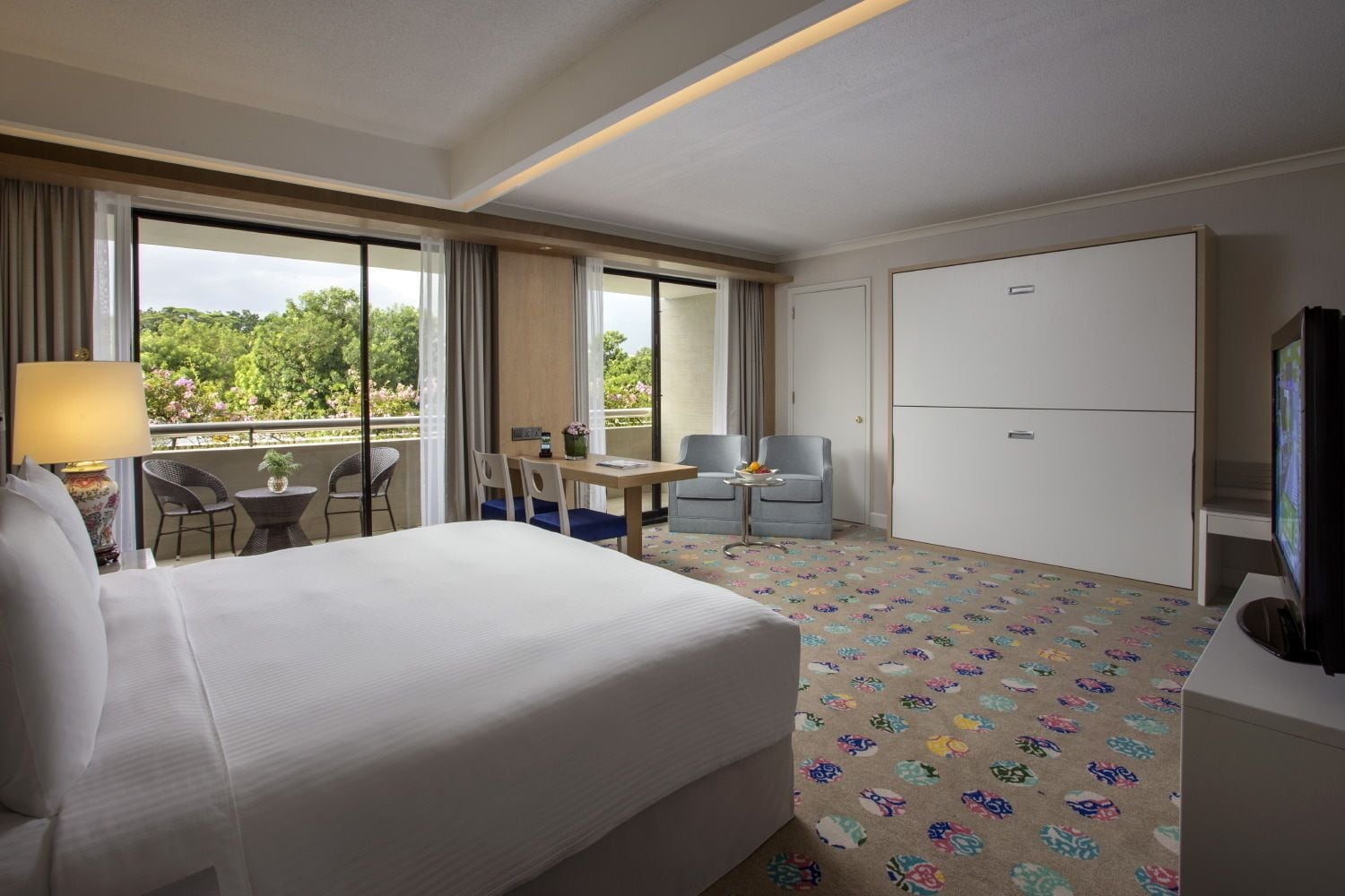 Une chambre d’hôtel avec un grand lit, un bureau avec deux chaises, une télévision et un balcon offrant une vue sur les arbres et les fleurs.