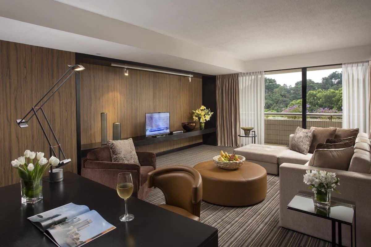 A modern hotel room with a sofa, armchair, and ottoman, a TV, and a balcony with a view of greenery.