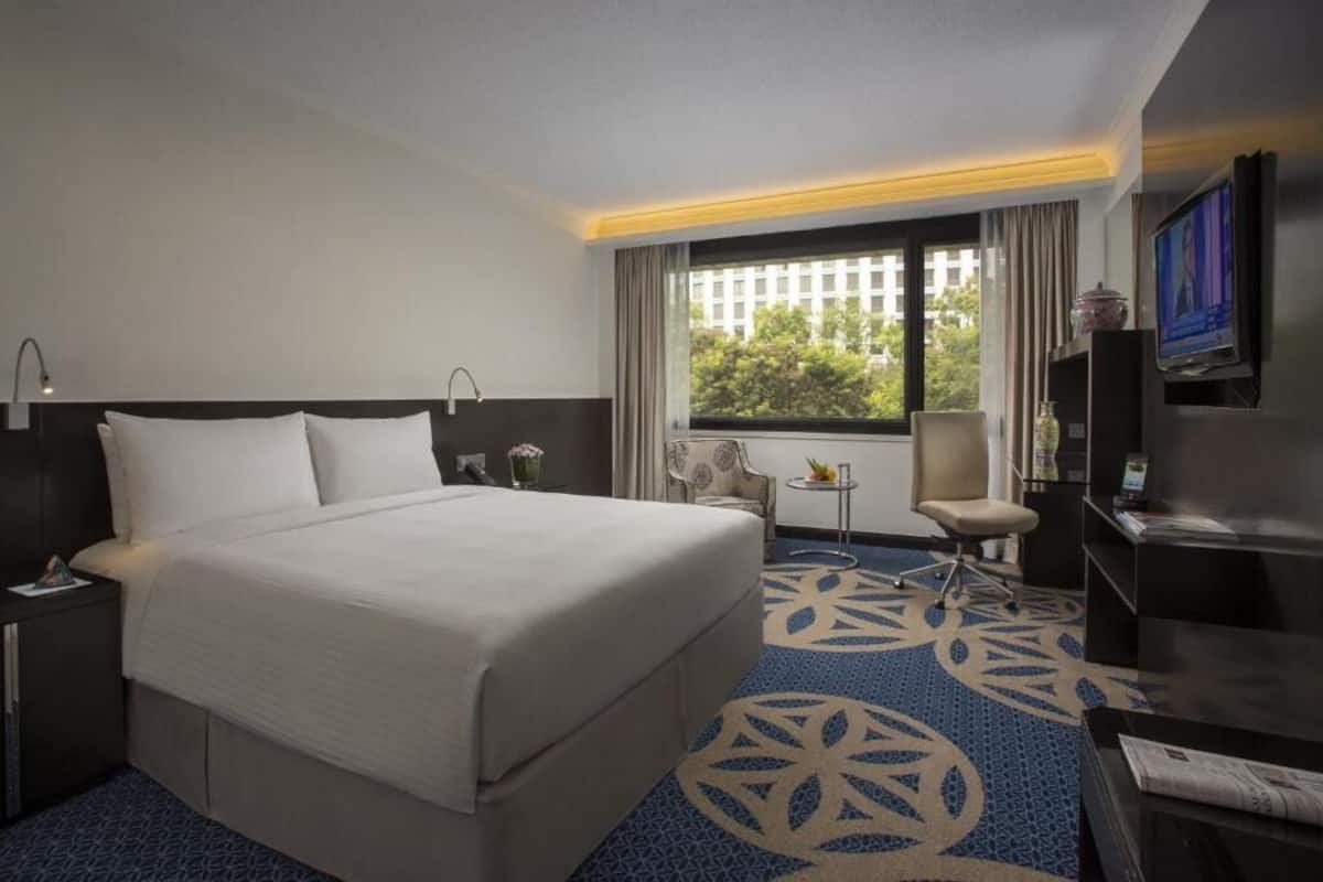 A hotel room with a large bed, a desk with a chair, a TV, and a window with a view of buildings and trees.