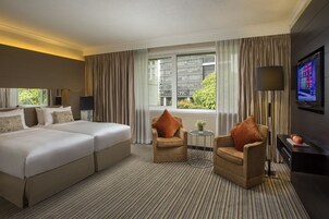 A hotel room with a large bed, two armchairs, a small table, a TV, and a window with curtains.