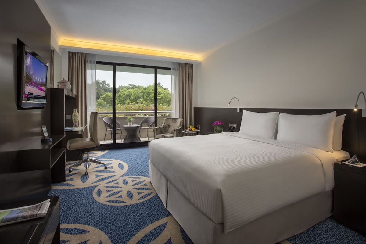 A hotel room with a large bed, a desk with a TV, a balcony with chairs, and a view of greenery.