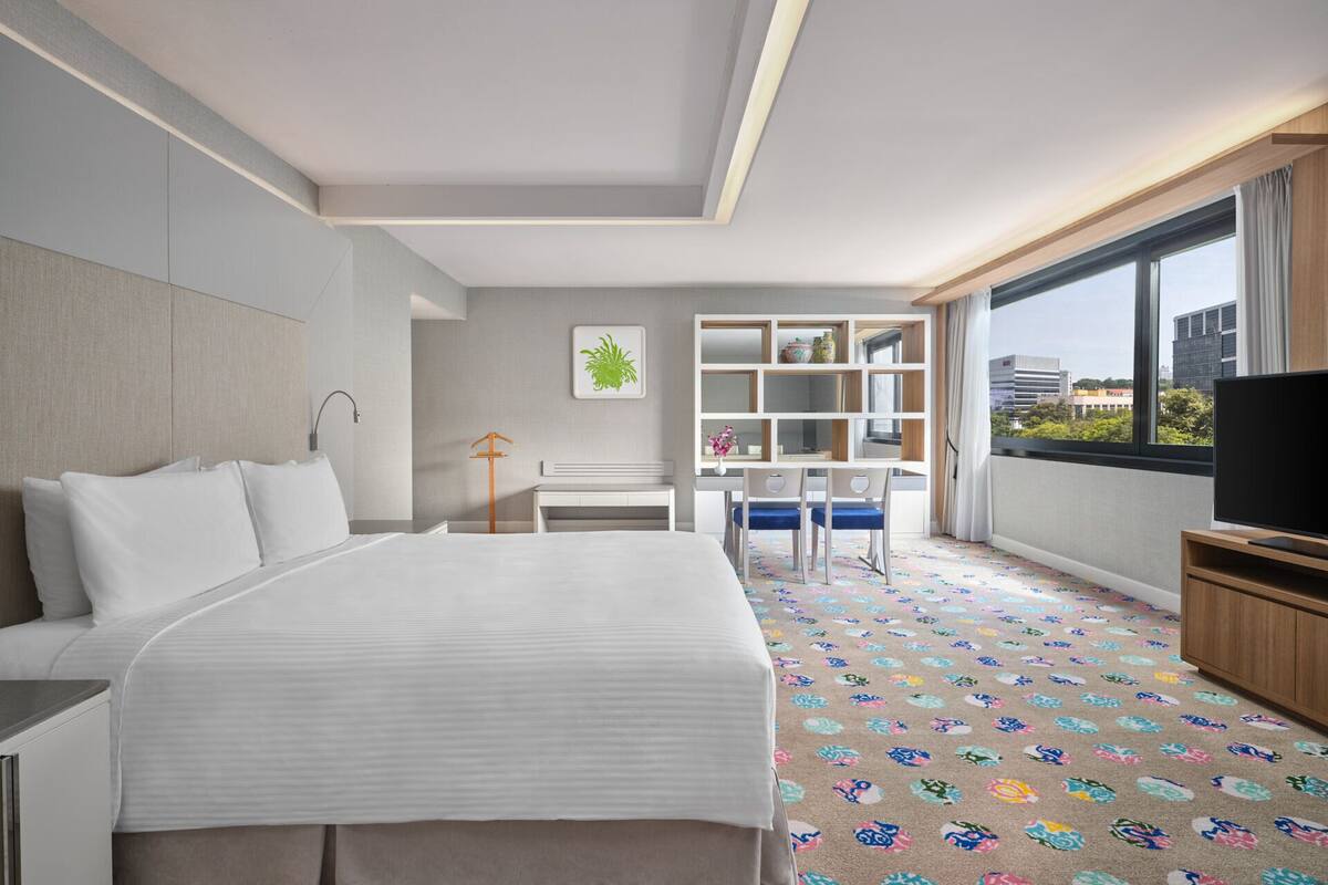 A hotel room with a large bed, a desk with a chair, a television, and a window with a city view.