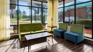 A room with a green sofa, blue chairs, a small table, and large windows offering a view of a building with a green roof and a parking lot. - Days Inn by Wyndham Petersburg/South Fort Lee (Petersburg)