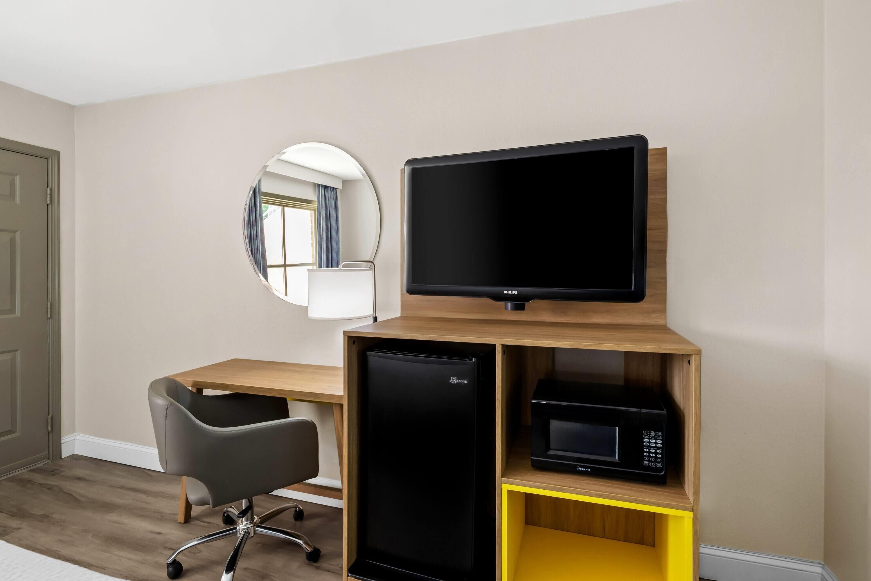 A compact hotel room with a wooden desk, a black refrigerator, a microwave, a black office chair, and a round mirror on the wall.