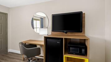 A compact hotel room with a wooden desk, a black refrigerator, a microwave, a black office chair, and a round mirror on the wall.