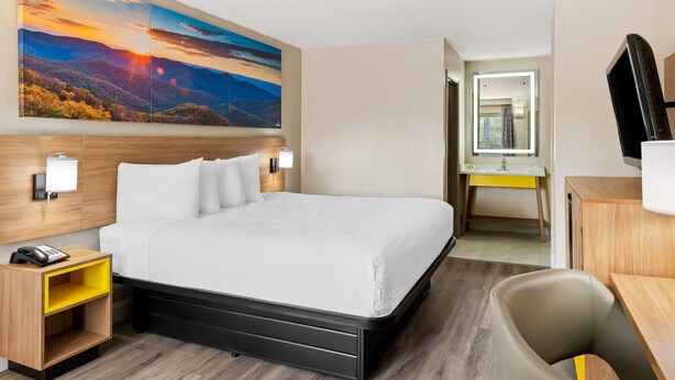 A hotel room with a large bed, a desk, a chair, a bedside table, a telephone, a mirror, and a painting of a sunset landscape. - Days Inn by Wyndham Petersburg/South Fort Lee (Petersburg)