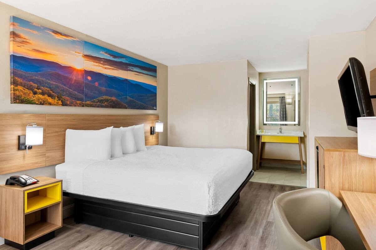 Une chambre d’hôtel avec un grand lit, un bureau, une chaise, une table de chevet, un téléphone, un miroir et un tableau représentant un paysage au coucher du soleil.