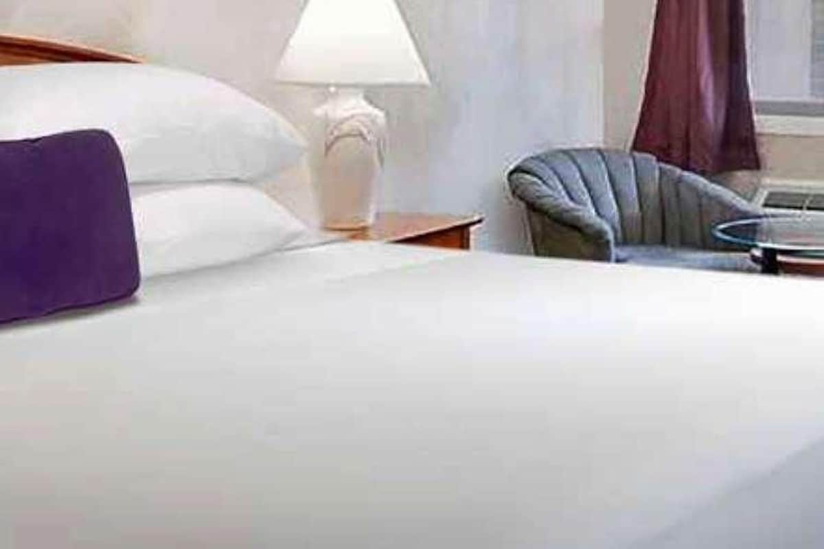 Una cama bien hecha con sábanas blancas y un cojín morado, un cabecero de madera, una lámpara de noche y una silla con una mesita redonda.