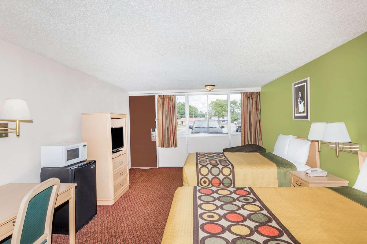 A hotel room with a desk, two beds, a TV, a refrigerator, a window with curtains, and a view of the outside.