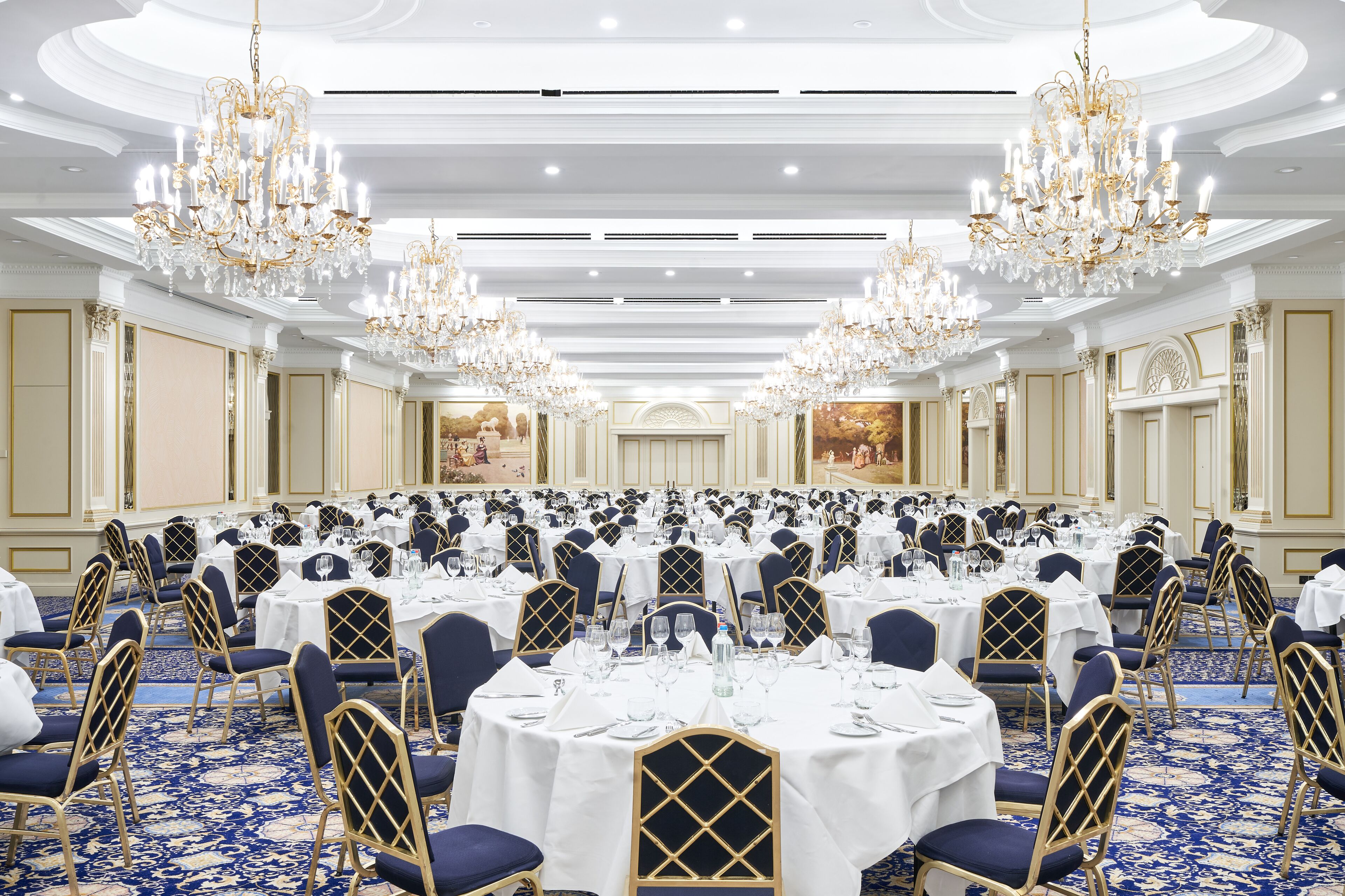 A large banquet hall with round tables, blue chairs, and chandeliers.