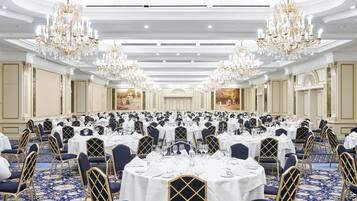 A large banquet hall with round tables, blue chairs, and chandeliers.