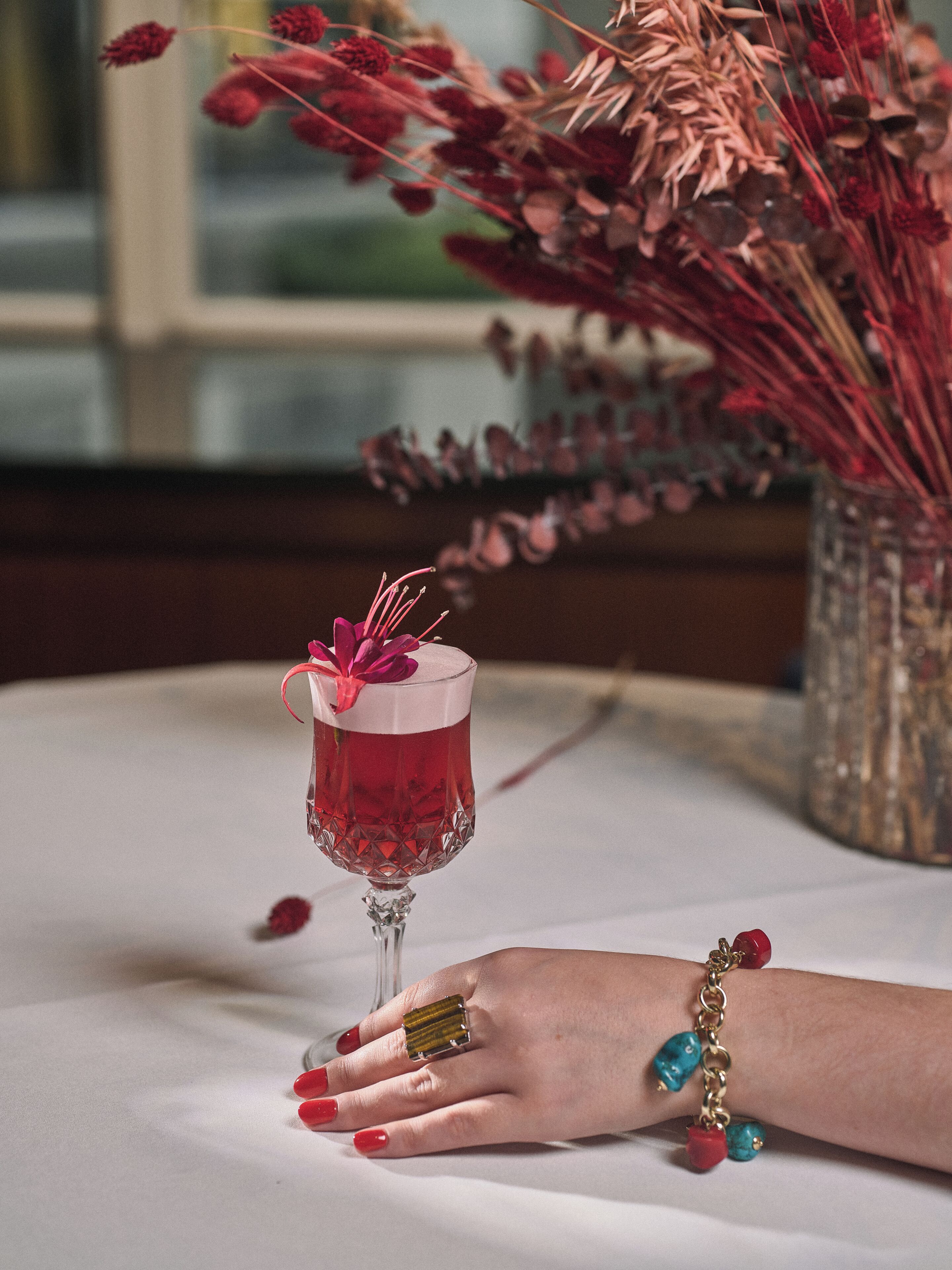 Une main portant une bague et un bracelet, posée sur une table avec un verre d’une boisson rouge et des fleurs séchées.