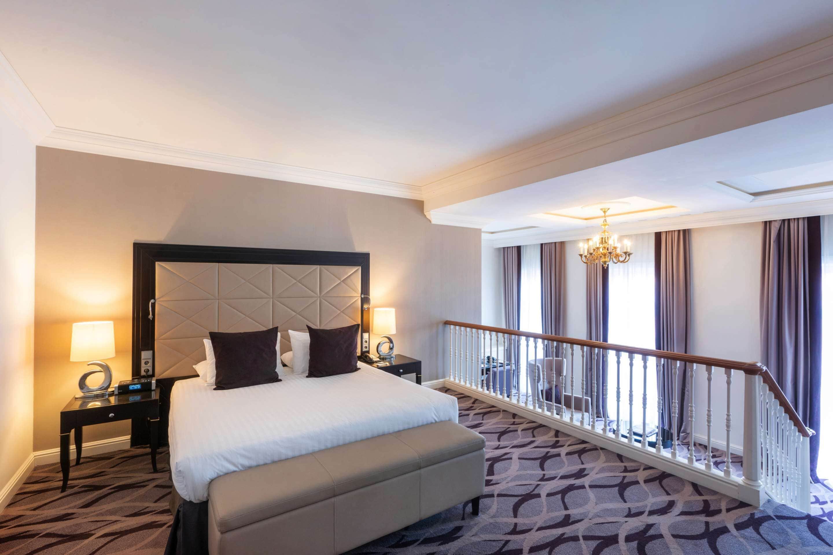 A hotel room with a large bed, a nightstand with a lamp, a modern chandelier, and a balcony with a railing.