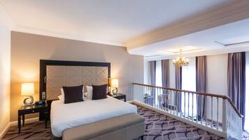 Une chambre d’hôtel avec un grand lit, une table de chevet avec une lampe, un lustre moderne et un balcon avec une balustrade.