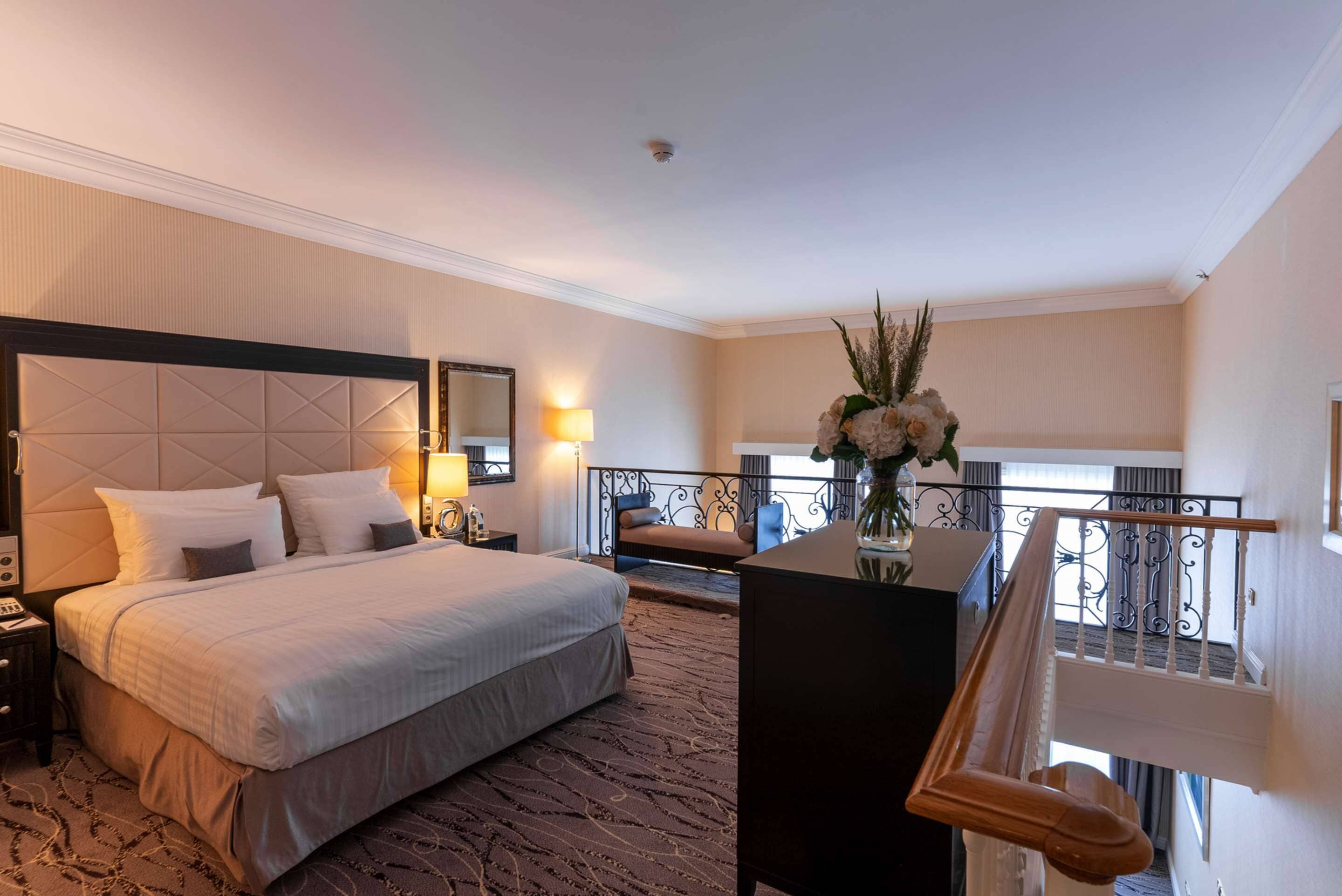 Habitación de hotel con una cama grande, un escritorio con un florero, un balcón con barandilla y una escalera.