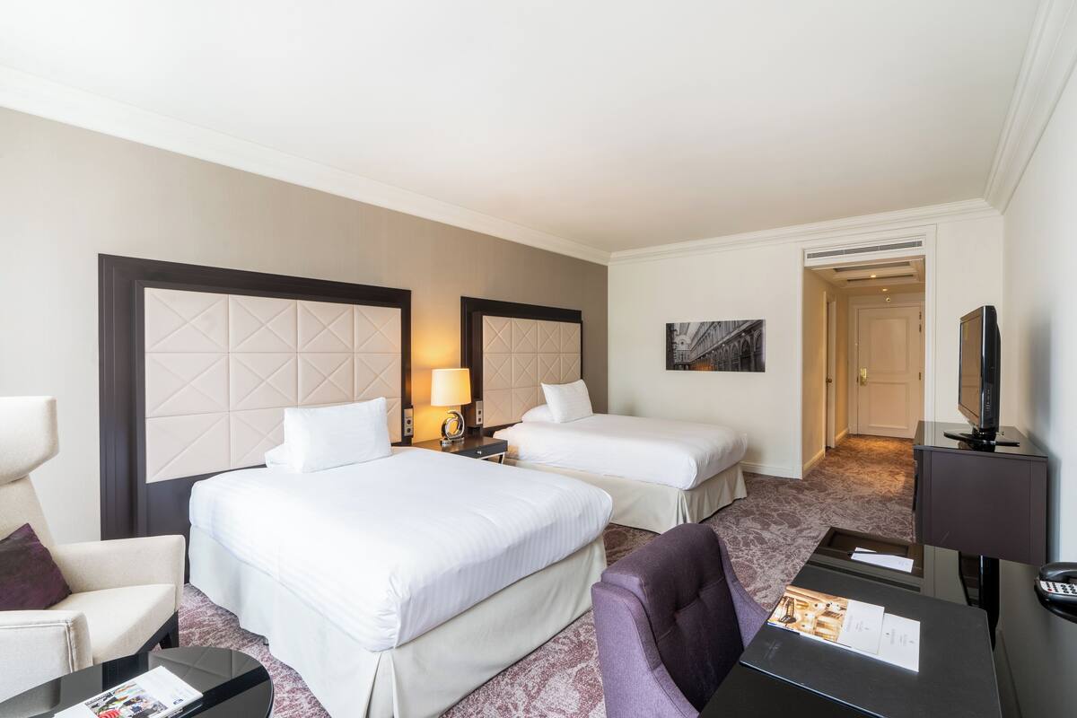 A hotel room with two beds, a desk, a chair, a TV, and a door leading to another room.