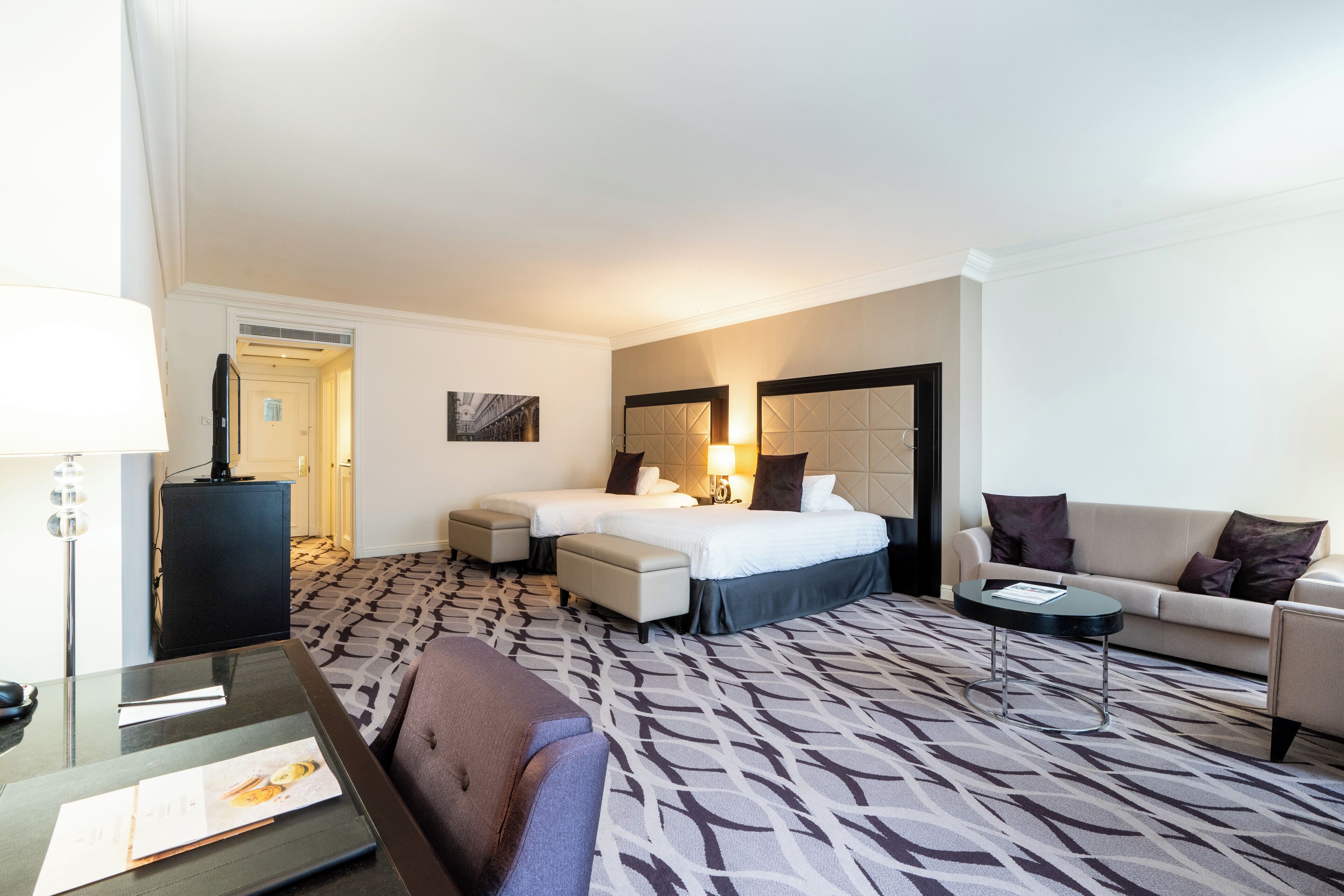 Quarto de hotel com cama, duas mesas de cabeceira, sofá, escrivaninha e um tapete estampado.