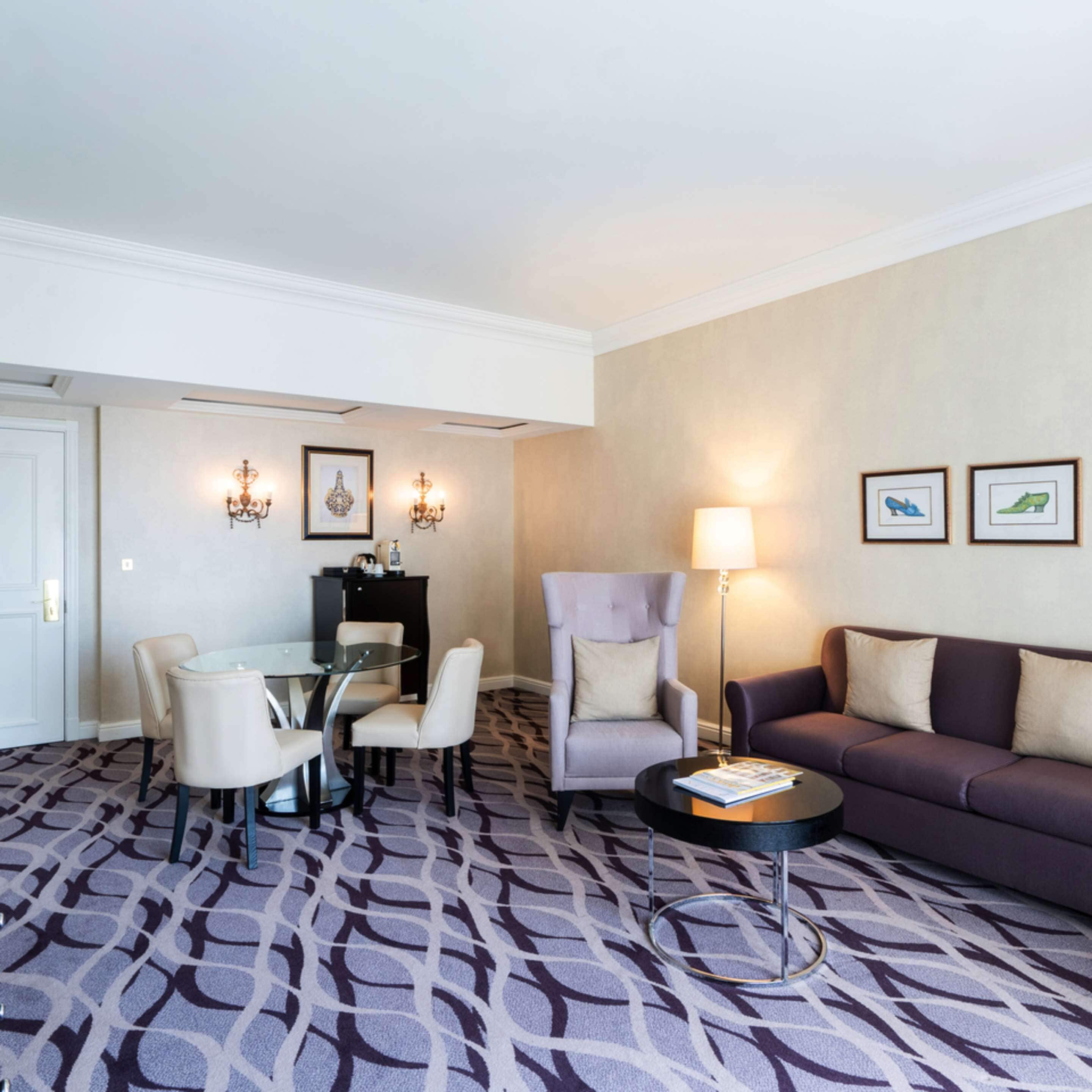 A hotel room with a dining area, a sofa, a lamp, and framed pictures on the wall.