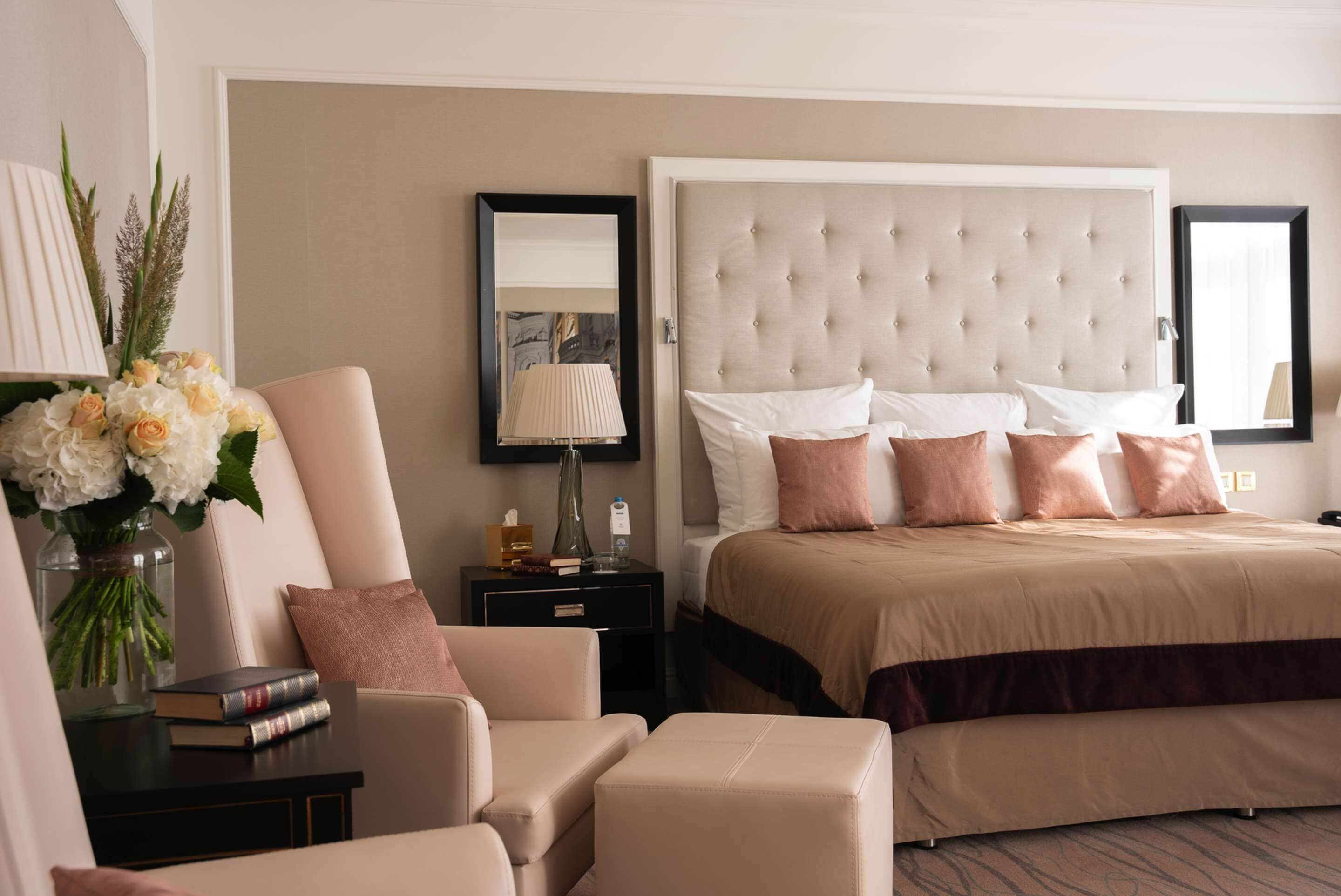 Une chambre d’hôtel avec un grand lit, un coin salon comprenant un fauteuil et une petite table, une table de chevet avec une lampe, et un vase avec des fleurs.