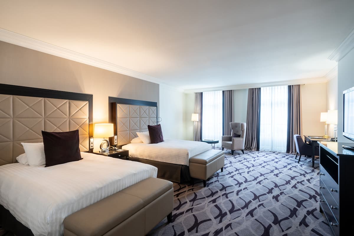 A hotel room with two beds, a desk, a chair, a TV, and a large window with curtains.