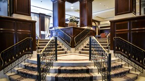 A grand staircase with ornate metal railings and carpeted steps, leading to a lobby area with a large vase of flowers. - Steigenberger Icon Wiltcher's (Brussels)