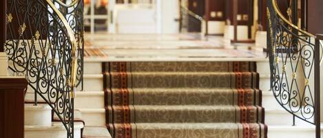 Una amplia escalera con barandales de metal ornamentados y alfombra estampada que conduce a un rellano en el segundo piso.
