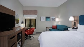 Una habitación de hotel con cama, escritorio con televisor, una silla roja, un sofá azul y obras de arte en las paredes.