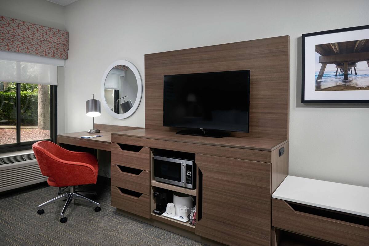 Hotellihuoneessa on puinen työpöytä, litteänäyttöinen televisio, punainen toimistotuoli, kahvinkeitin ja seinällä oleva kehystetty kuva.