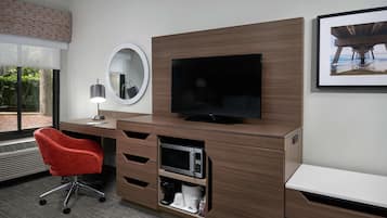 Habitación de hotel con un escritorio de madera, un televisor de pantalla plana, una silla de oficina roja, una cafetera y un cuadro en la pared.