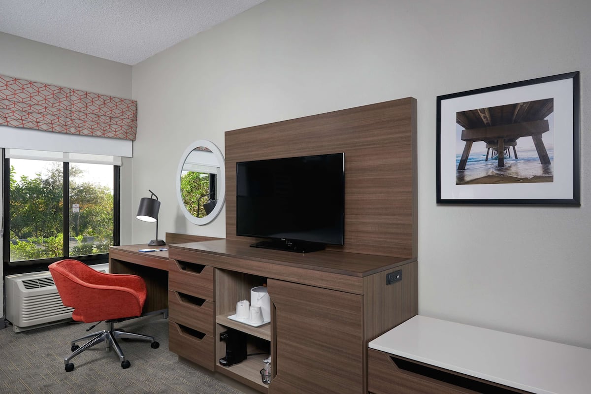 Hotellihuone, jossa on puinen työpöytä, litteänäyttöinen televisio, punainen toimistotuoli, suuri ikkuna, josta on näkymä vehreään maisemaan, ja seinällä oleva kehystetty taulu.