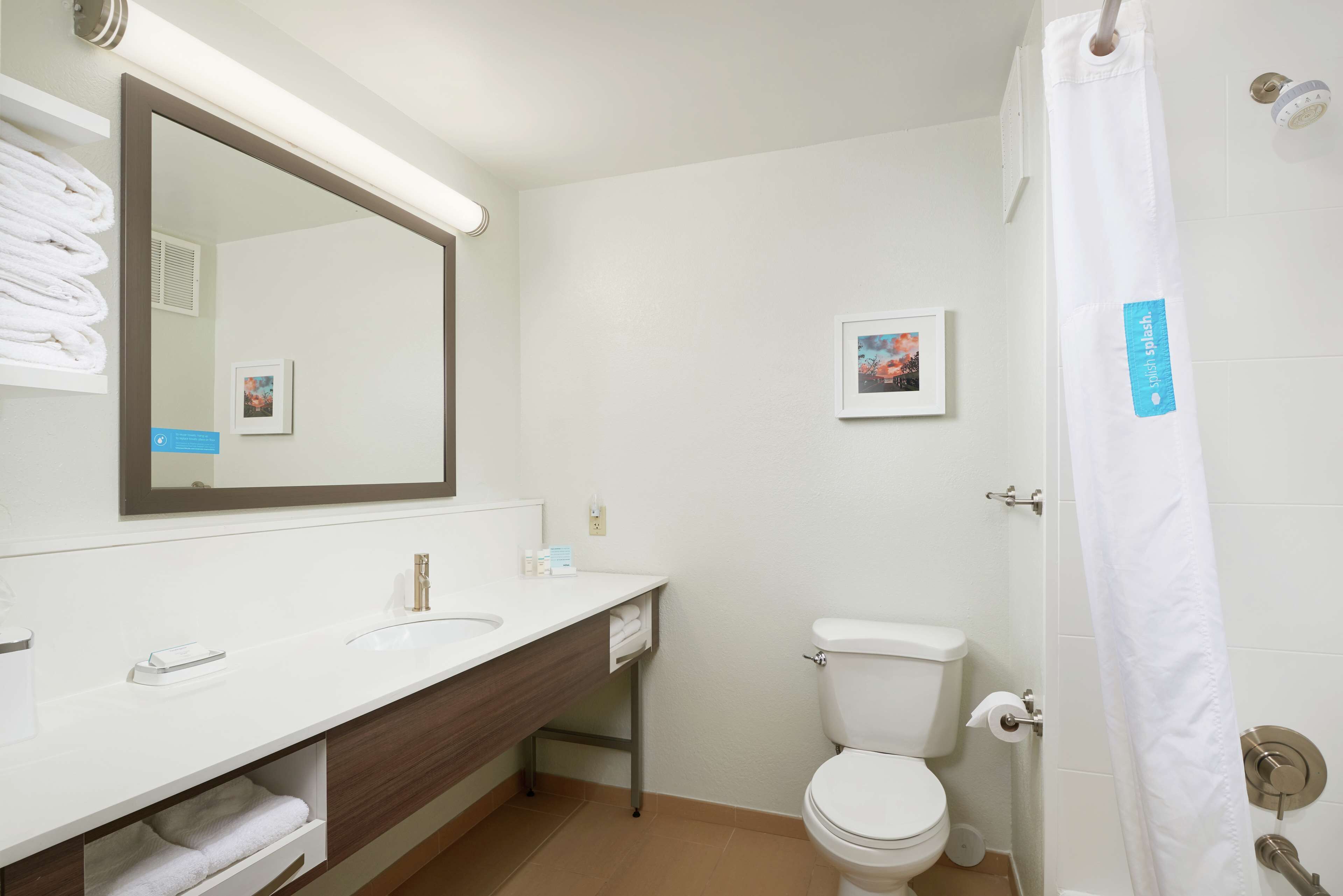 Ein modernes Badezimmer mit Waschbecken, Spiegel und Toilette. Ein Handtuchhalter mit gefalteten Handtüchern, ein gerahmtes Bild an der Wand und ein Duschvorhang mit Etikett.