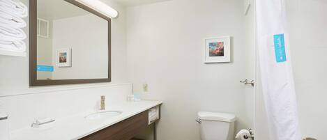 A modern bathroom with a white sink, mirror, and toilet. There is a towel rack with folded towels, a framed picture on the wall, and a shower curtain with a label.