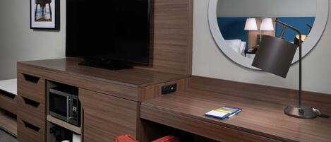 A hotel room with a flat-screen TV, a round mirror, a desk with a lamp, a red chair, and a microwave.